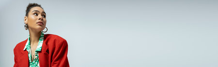 african american woman in stylish red blazer and hoop earrings looking away on grey backdrop, bannerの写真素材