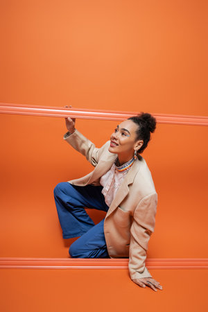 cheerful african american fashion model in stylish outfit squatting out of orange frameworkの写真素材