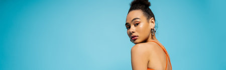 portrait of african american woman with hoop earrings looking at camera on blue backdrop, banner