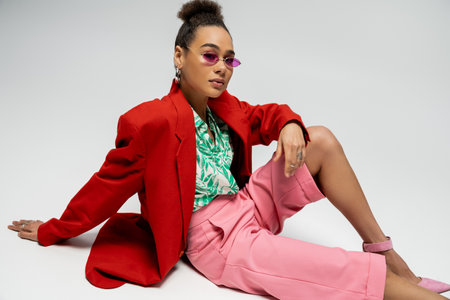 confident african american woman in stylish attire and pink sunglasses sitting on grey backdropの写真素材