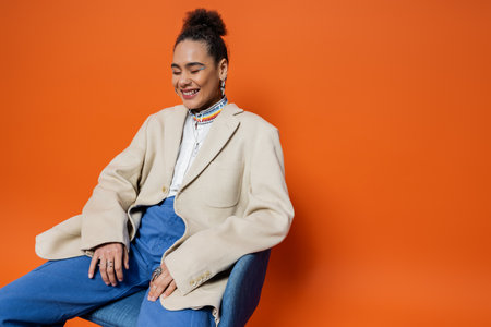 cheerful fashion model in trendy attire sitting on chair and smiling sincerely with closed eyesの写真素材