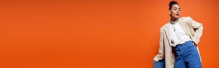 pretty african american model in beige blazer with vibrant makeup standing near blue chair, bannerの写真素材