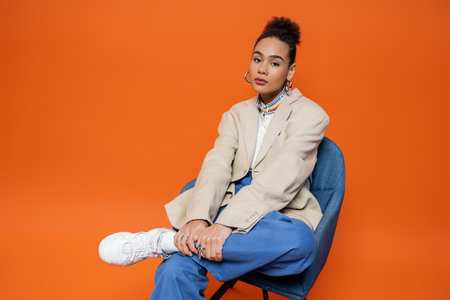 young woman in stylish street outfit with vivid makeup bun and accessories sitting on blue chairの写真素材