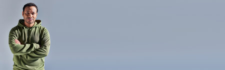 cheerful young indian man in khaki hoodie with hands crossed on chest on yellow background, bannerの写真素材