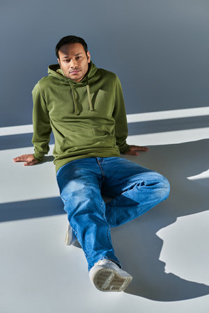 young indian man in casual urban attire slightly smiling sitting on floor and looking at camera