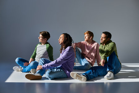 four young men in casual outfits sitting on floor on grey background and looking away, diversityの写真素材