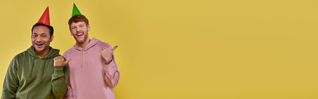 two young handsome men in birthday hats pointing thumbs aside and smiling at camera, birthdayの写真素材