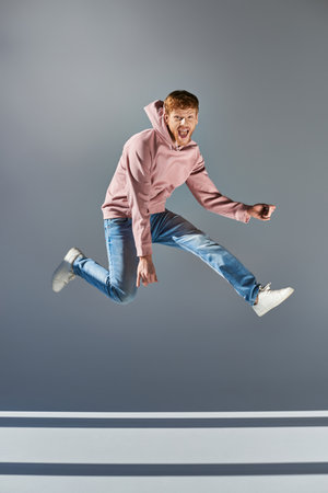 cheerful young man in casual clothing jumping and playing invisible guitar on grey backdropの写真素材