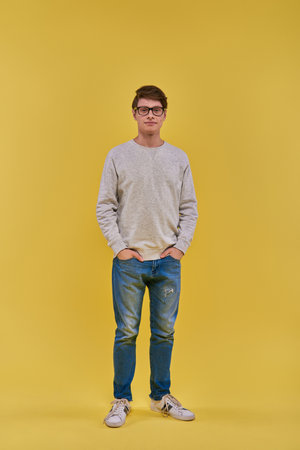 young handsome man in sweatshirt and jeans standing still with hands in pockets looking at cameraの写真素材