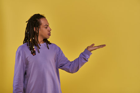 confused african american man in purple sweatshirt with dreadlocks holding hand empty palm upの写真素材