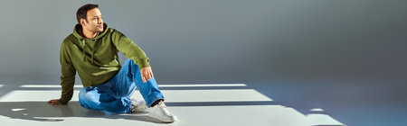 young indian man in trendy khaki hoodie sitting on floor looking away on grey backdrop, bannerの写真素材