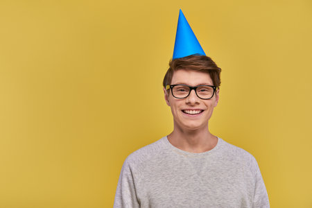 young cheerful man in white sweatshirt and birthday hat smiling at camera on yellow backdropの写真素材