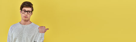 young man in simple wear and glasses pointing finger aside on left side on yellow backdrop, bannerの写真素材