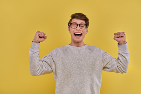 emotional happy man in casual street outfit raising fists up and closing his eyes on yellow backdropの写真素材
