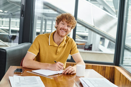 cheerful red haired man smiling and looking at camera while working on his papers, coworking conceptの写真素材