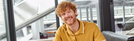 happy red haired man smiling and looking at camera with window backdrop, coworking concept, bannerの写真素材