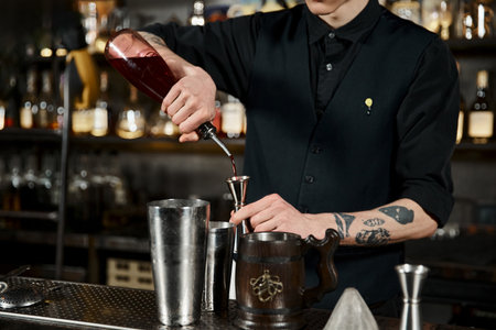 cropped view of bartender pouring alcohol drink into jigger near cocktail shakers and wooden mugのeditorial素材