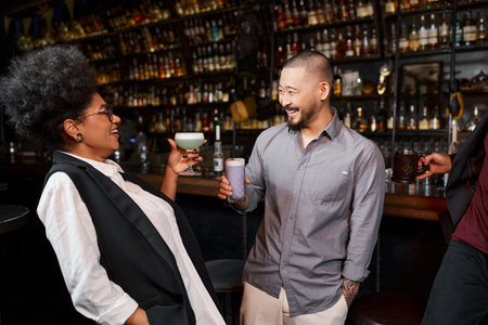 cheerful asian man and african american woman with drinks smiling at each other in cocktail barの写真素材