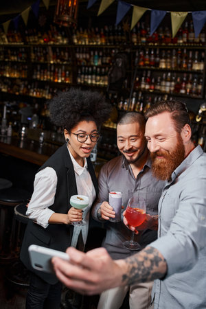 tattooed bearded man taking selfie on smartphone with smiling multiethnic colleagues in cocktail barの写真素材