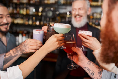 joyful multiethnic workmates clinking glasses with cocktails in bar after work, blurred backgroundの写真素材