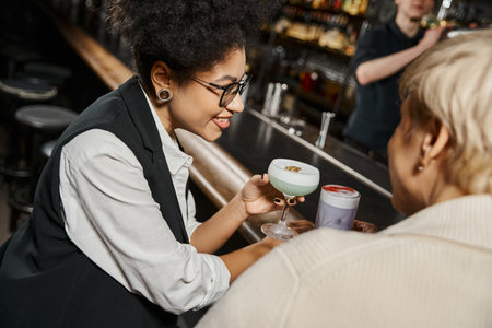 african american woman with cocktail glass smiling near female workmate in bar on blurred foregroundの写真素材