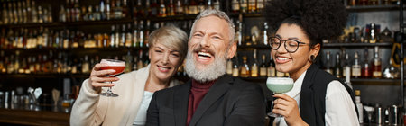 happy multiethnic women holding cocktail glasses near bearded colleague laughing in bar, bannerの写真素材