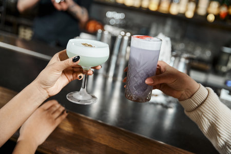 cropped view of multiethnic female colleagues holding delicious cocktails in bar after work