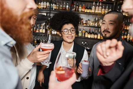 positive african american woman holding cocktail and smiling near multiethnic work friends in barの写真素材