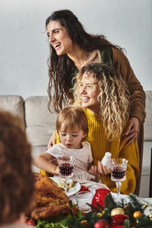 young lgbt couple with their daughter sitting at Christmas table surrounded by their relativesの写真素材