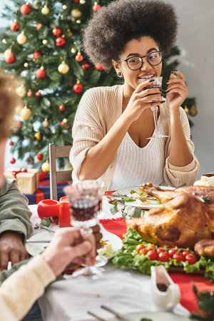 cheerful african american woman talking to her relatives drinking wine with Christmas tree backdropの写真素材