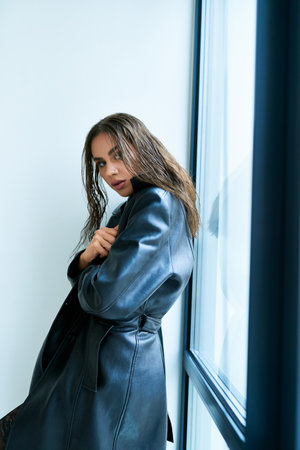 attractive woman with brunette long hair posing in black leather coat near window at home, seductiveの写真素材