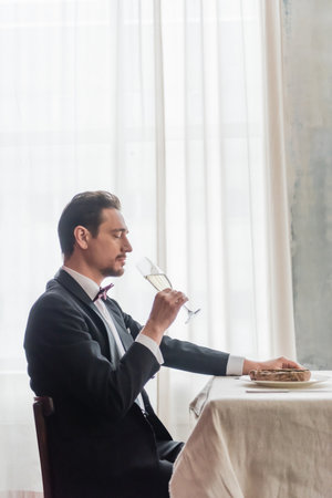 handsome man in tuxedo enjoying taste of champagne and sitting at table with beef steak on plateの写真素材