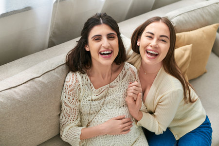 joyful lgbt couple sitting on sofa and smiling cheerfully at camera, in vitro fertilisation conceptの写真素材