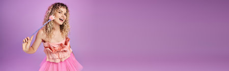 cheerful curly woman in pink dress with magic wand smiling at camera, tooth fairy concept, bannerの写真素材
