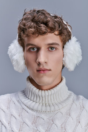 portrait of young man with snow on wavy hair posing in warm earmuffs and white sweater on greyの写真素材