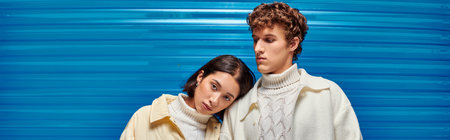 young asian model leaning on shoulder of stylish man on blue plastic backdrop, winter trend, banner