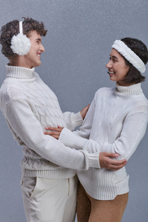 cheerful interracial couple in cozy winter attire looking at each other on grey background with snowの写真素材