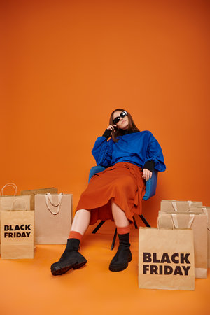 tired woman in stylish sunglasses sitting on armchair near black friday shopping bags on orangeの写真素材