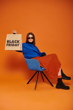 woman in sunglasses and autumnal attire holding black friday shopping bag and sitting on armchairの写真素材