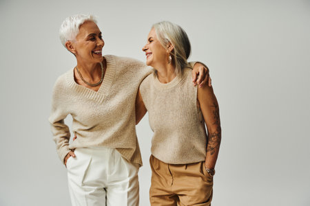 fashionable senior female friends smiling at each other while posing with hands in pockets on greyの写真素材