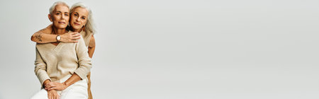 senior tattooed woman embracing smiling female friend sitting on grey,  lifelong friends, bannerの写真素材