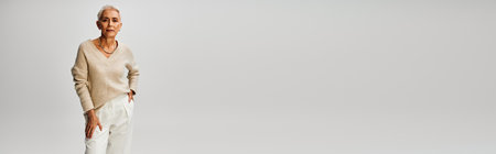 trendy senior woman in knitted jumper and pants standing with hand in pocket on grey, bannerの写真素材