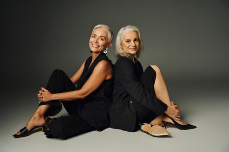 joyful and fashionable senior women in black clothes sitting back to back on grey backdrop, eleganceの写真素材