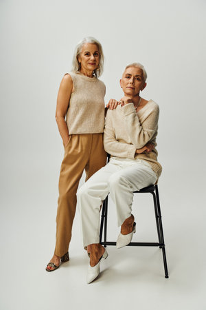 elegant senior woman sitting on chair and looking at camera near smiling female friend on greyの写真素材