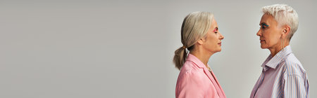 side view of trendy mature women in blue and pink casual attire posing on grey backdrop, banner