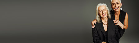 pleased and trendy senior models in black casual attire smiling at camera on grey backdrop, bannerの写真素材