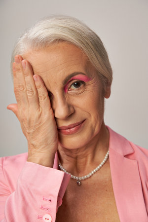 portrait of elegant mature lady in pearl necklace and pink blazer obscuring face with hand on greyの写真素材
