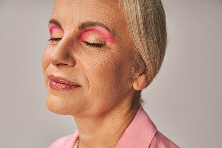 close-up portrait of stylish senior lady with natural makeup smiling with closed eyes on greyの写真素材