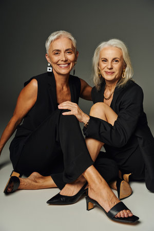 charming elegant senior models in black wear sitting and smiling at camera on grey, stylish agingの写真素材