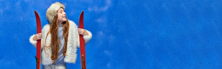winter joy, pleased preteen girl in faux fur jacket and hat holding red skis on turquoise, bannerの写真素材